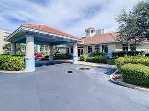 Ein Hotel mit Eingangsbereich, überdachter Auffahrt, Palmen und einem Gebäude mit rotem Ziegeldach.