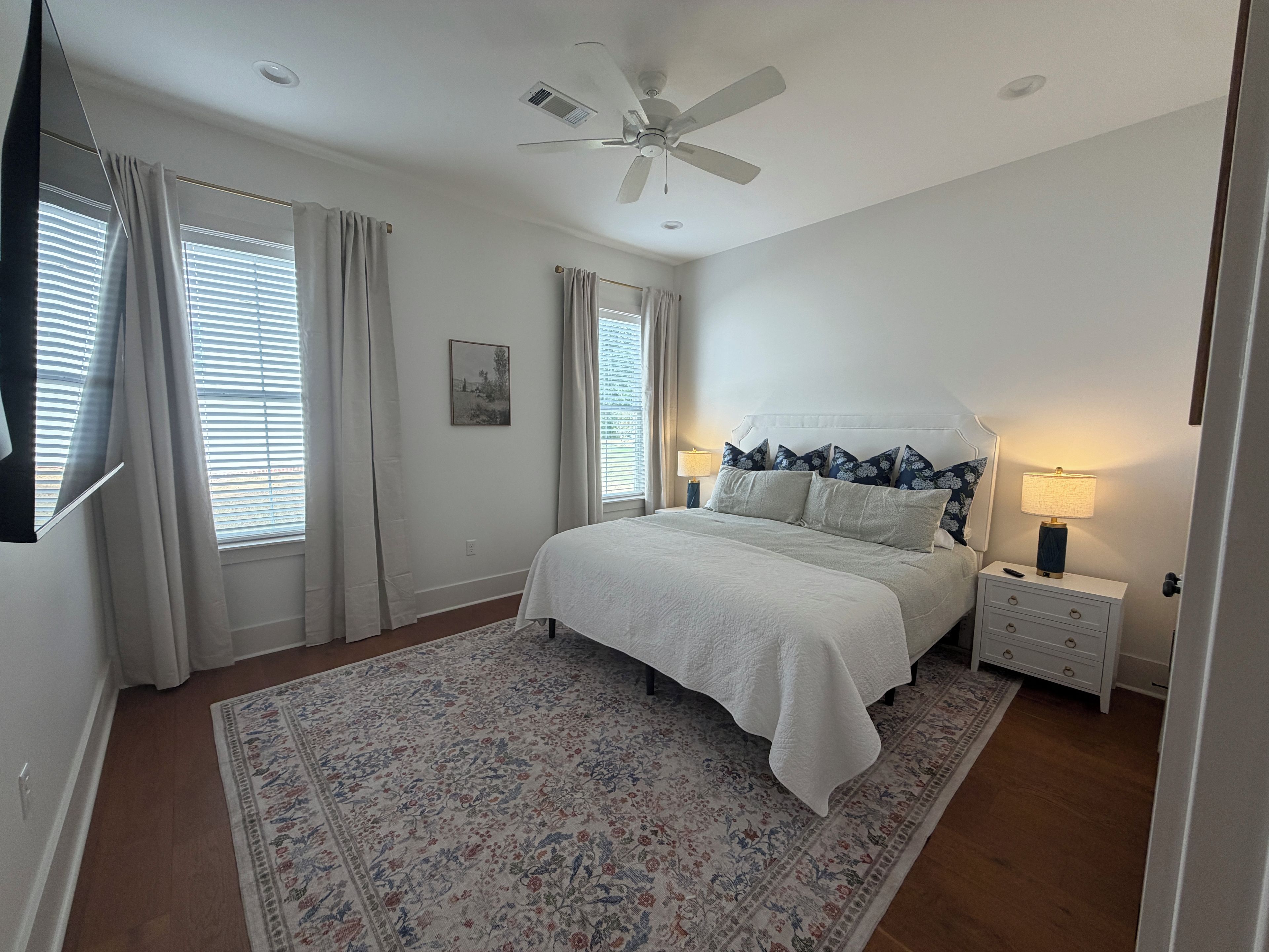 Une chambre à coucher avec un lit, des tables de chevet, un ventilateur de plafond et une fenêtre avec des rideaux.