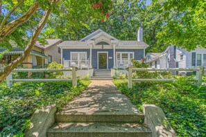 A two-story house with a front porch, a stone pathway leading to the entrance, and a white picket fence. - The Hamilton a Stylish Downtown Bungalow (High Point)