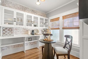 A kitchen with white cabinetry, a brick accent wall, a round dining table with chairs, and a window with blinds. - The Hamilton a Stylish Downtown Bungalow (High Point)