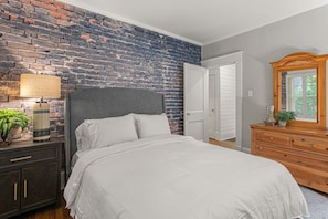 Ein Schlafzimmer mit Backsteinwand, einem Bett mit weißer Bettwäsche, einem Schminktisch, einer Lampe und einem Spiegel.