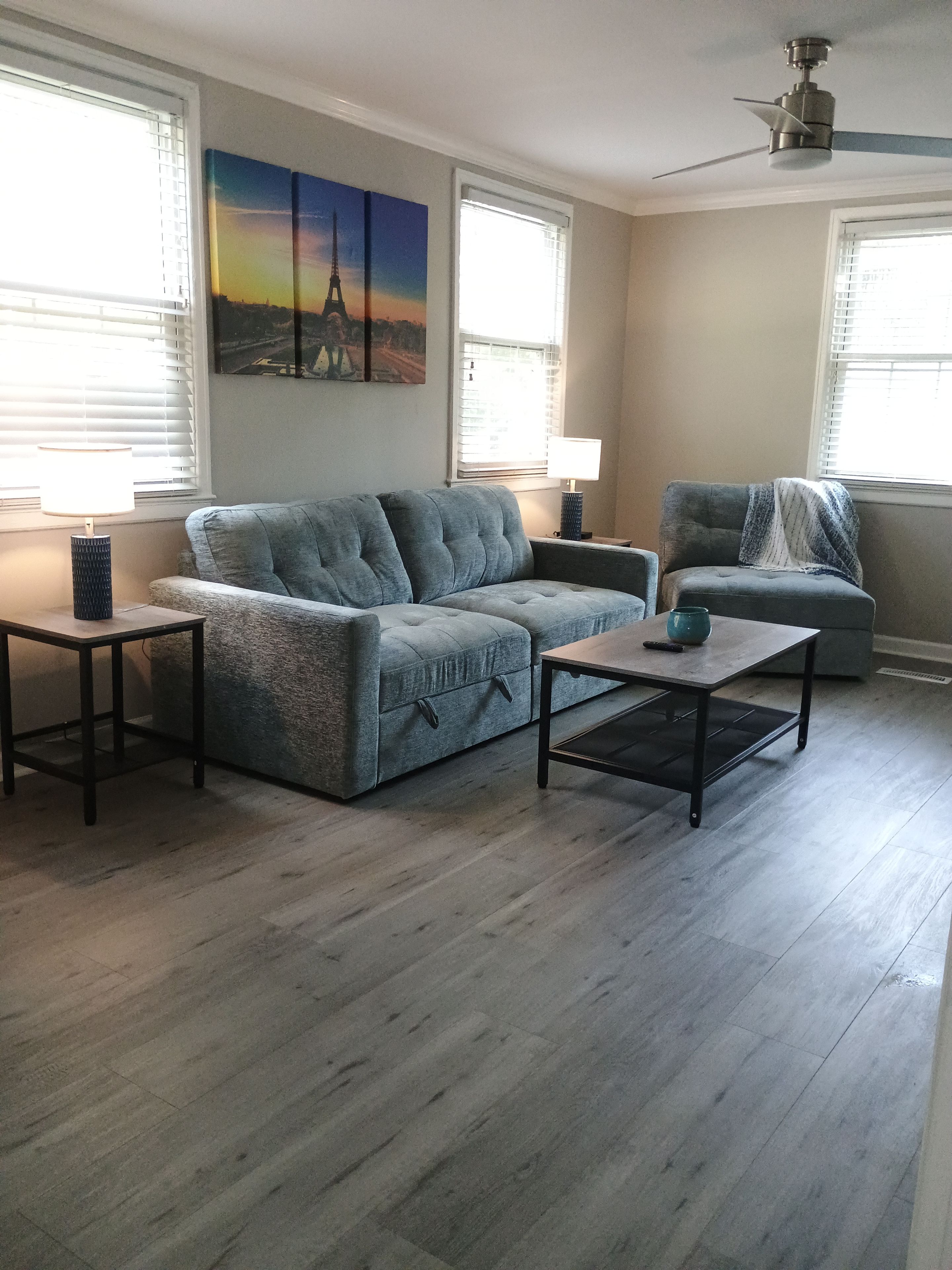 A living room with a sectional sofa, a coffee table, and a side table. There are two windows with blinds, a ceiling fan, and a painting of the Eiffel Tower.
