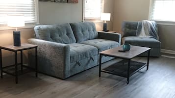 A living room with a sectional sofa, a coffee table, and a side table. There are two windows with blinds, a ceiling fan, and a painting of the Eiffel Tower.