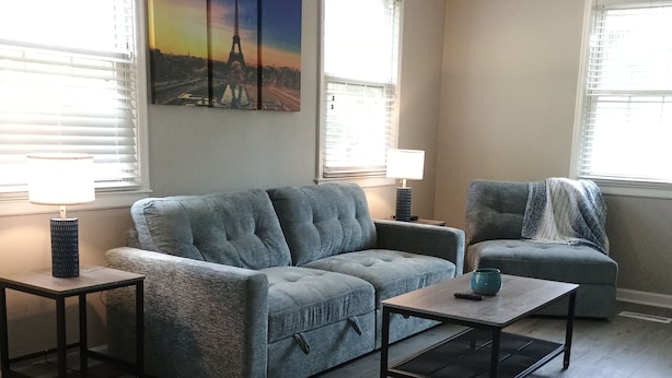 A living room with a sectional sofa, a coffee table, and a side table. There are two windows with blinds, a ceiling fan, and a painting of the Eiffel Tower. - Cozy home in Ardmore Winston-Salem pet friendly, close to hospital & University (Winston-Salem)