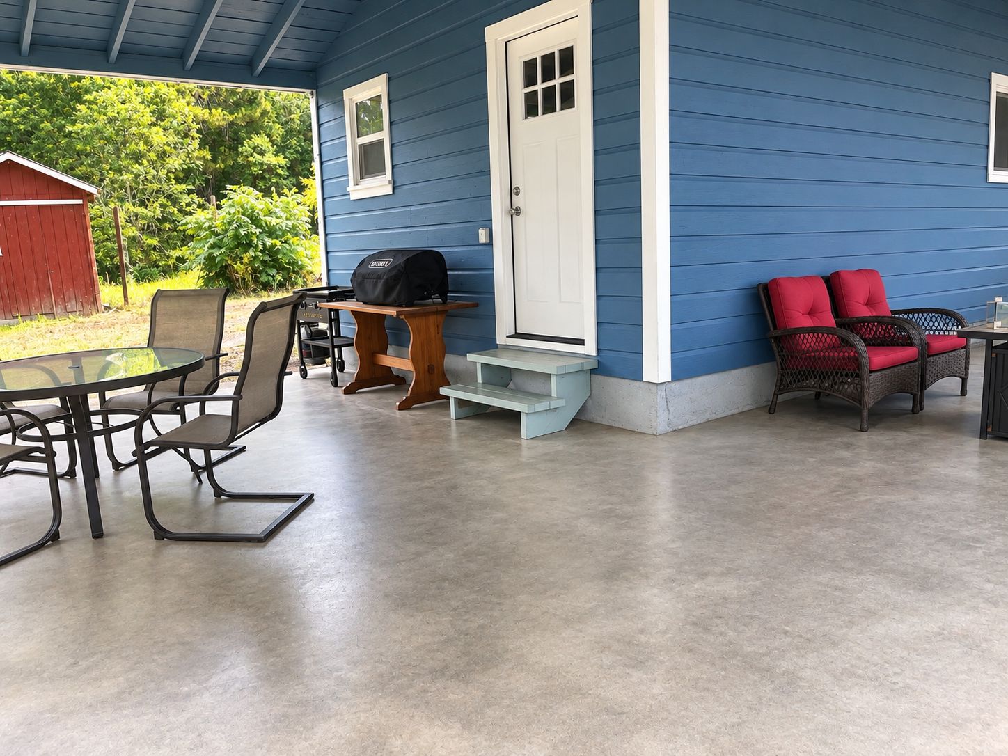 A covered patio area with a table and chairs, a grill, and a door leading to a blue house.