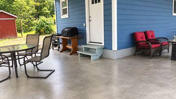 A covered patio area with a table and chairs, a grill, and a door leading to a blue house.
