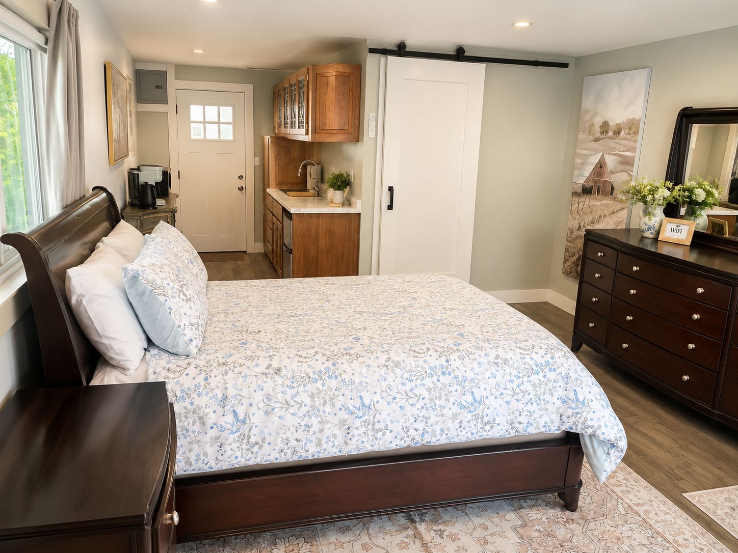 A bedroom with a large bed, a dresser, a nightstand, a desk, a window with curtains, a door, and a kitchen area with cabinets and a sink.