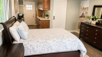 A bedroom with a large bed, a dresser, a nightstand, a desk, a window with curtains, a door, and a kitchen area with cabinets and a sink.