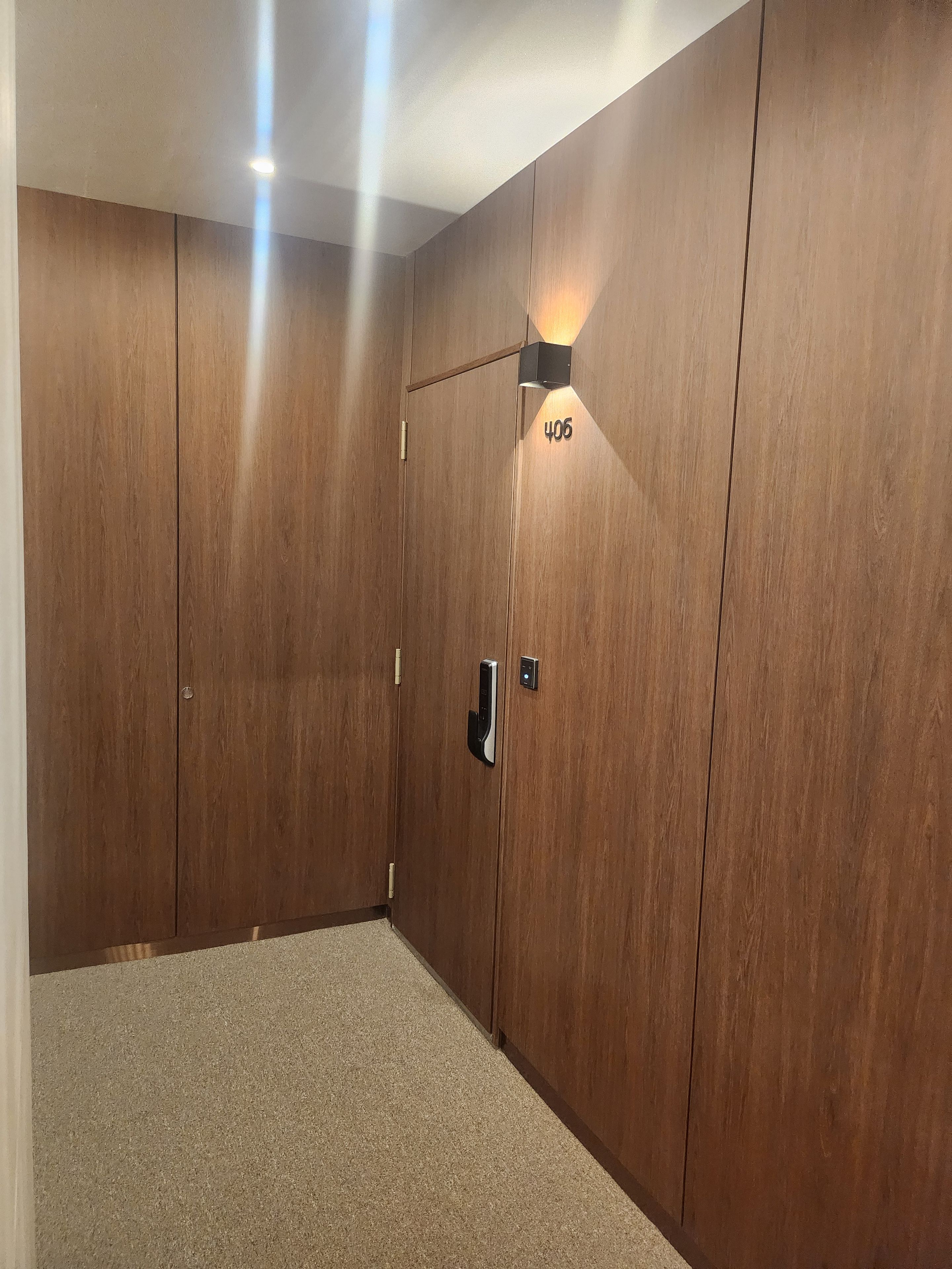 Una habitación con un armario de madera, una puerta con placa de número y una lámpara fijada a la pared.