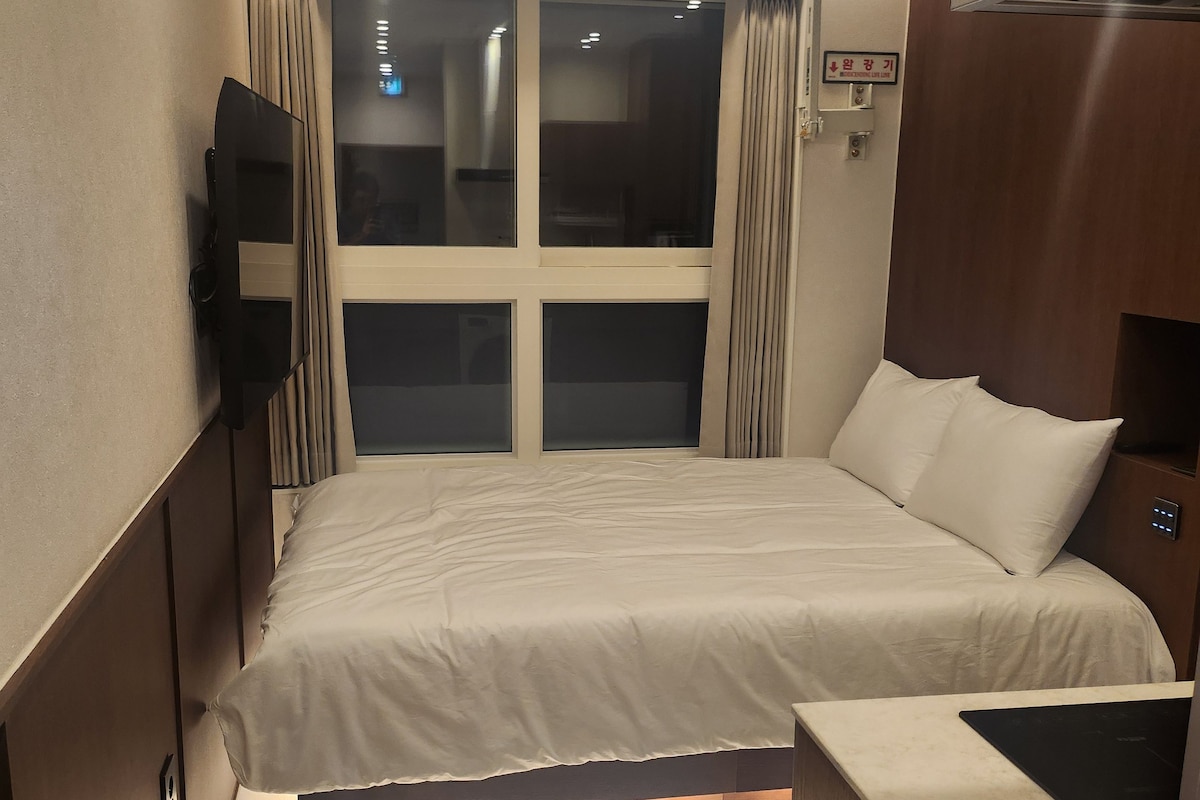 Habitación de hotel con cama, televisor montado en la pared, ventana con cortinas y cabecero de madera.