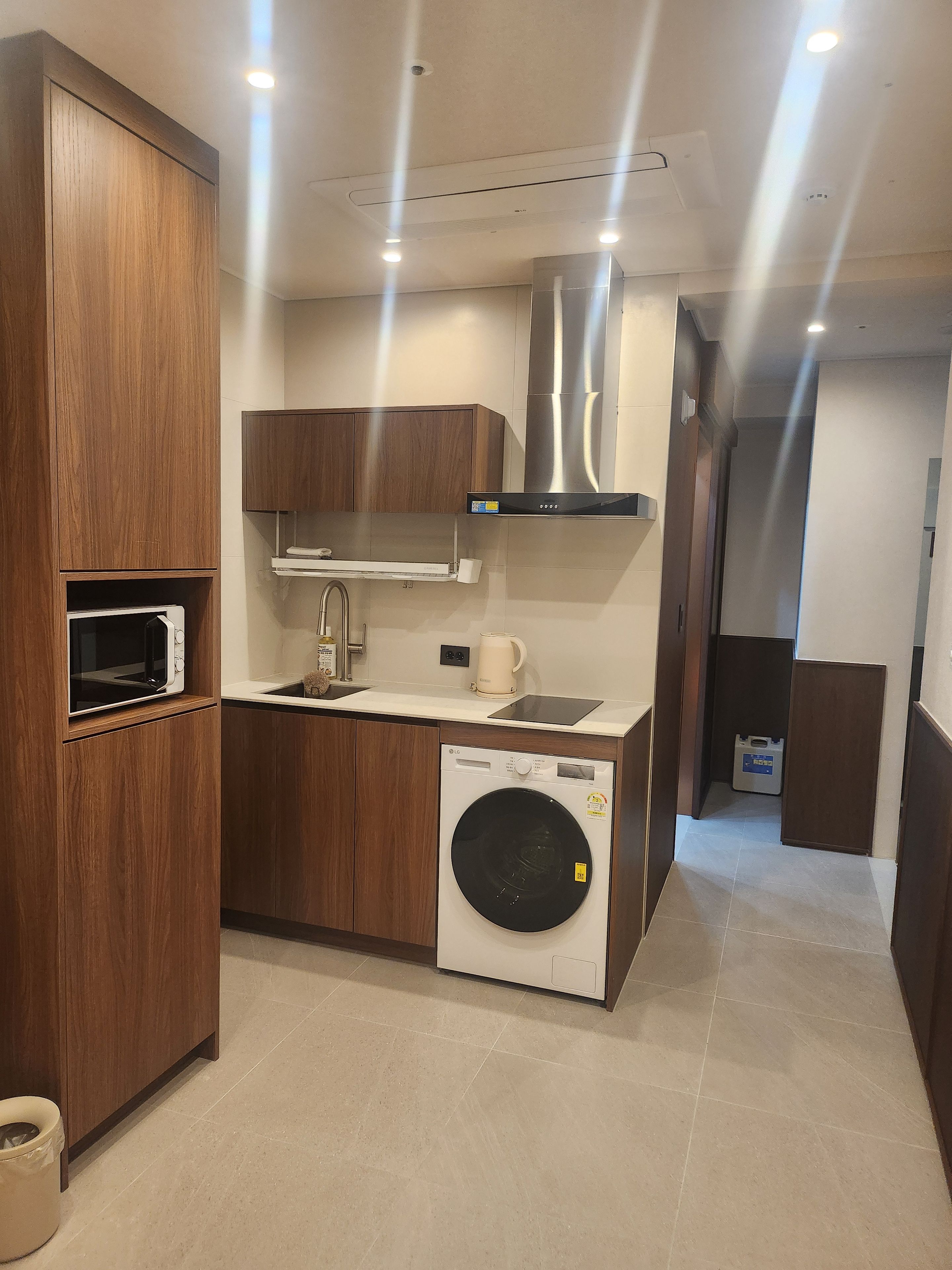 Cocina compacta con armarios de madera, microondas integrado, campana extractora de acero inoxidable, lavadora y un lavamanos.