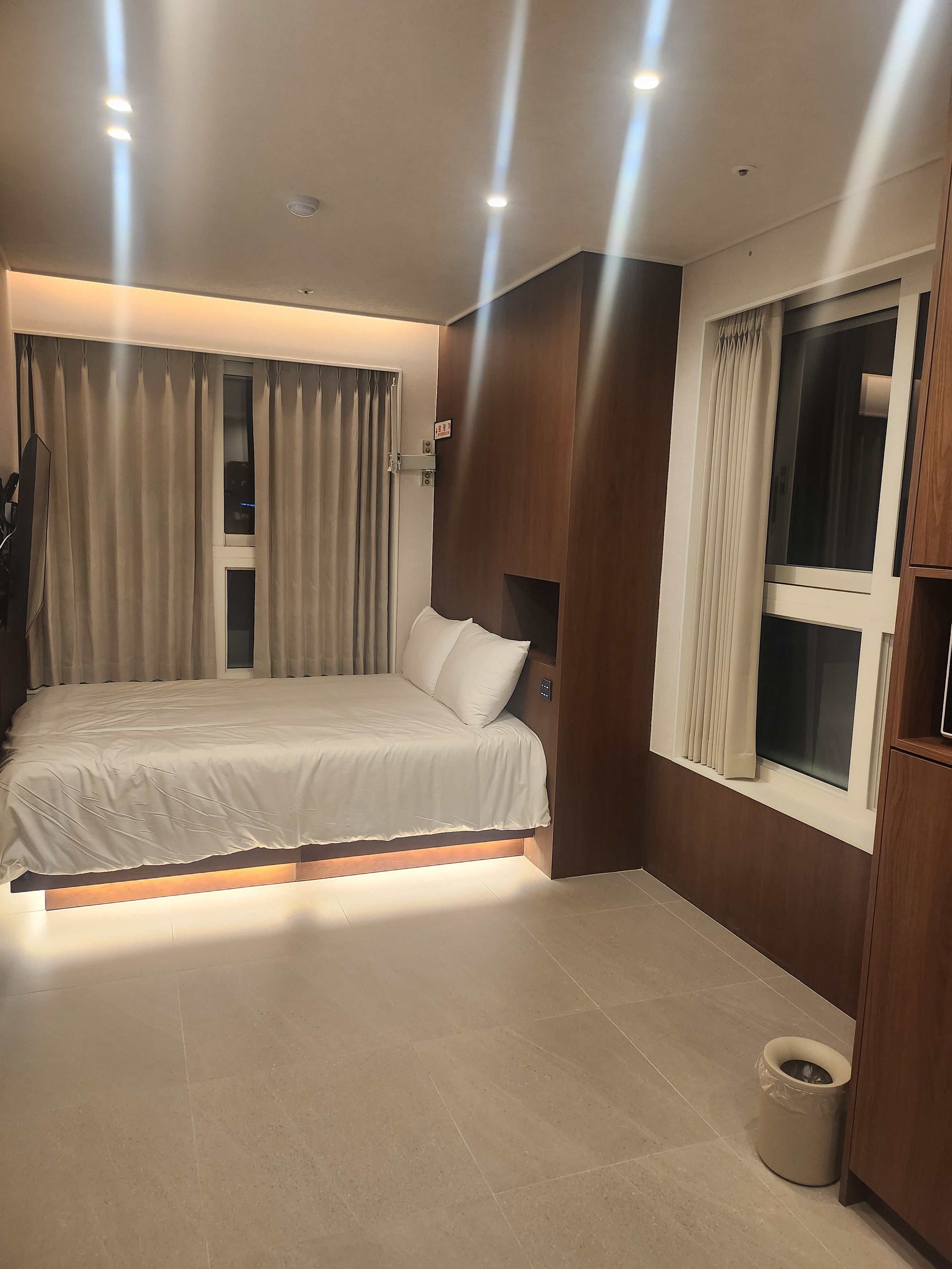 Un dormitorio con cama, cortinas, una ventana y un cubo de basura.