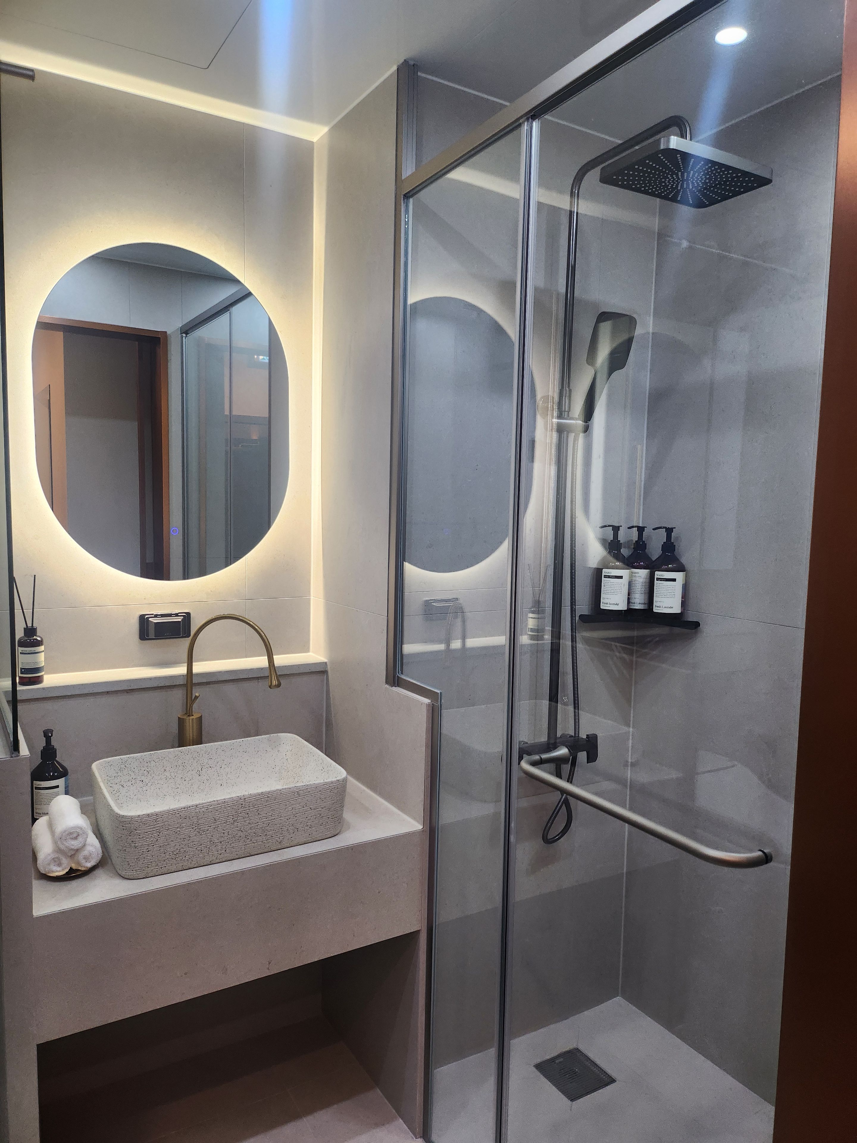 A modern bathroom with a glass-enclosed shower, a round mirror, a rectangular sink, and a small shelf with toiletries.