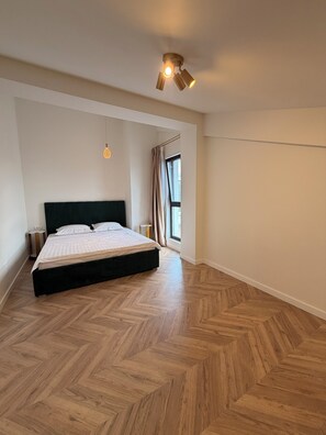 A bedroom with a large bed, a ceiling light fixture, a window with curtains, and a herringbone-patterned wooden floor. - Cozy 1-bedroom apartment in northside Bucharest with AC and terrace (București)