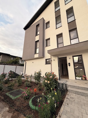 A modern building with a flat facade, multiple windows, and a small garden area with various flowers. - Cozy 1-bedroom apartment in northside Bucharest with AC and terrace (București)