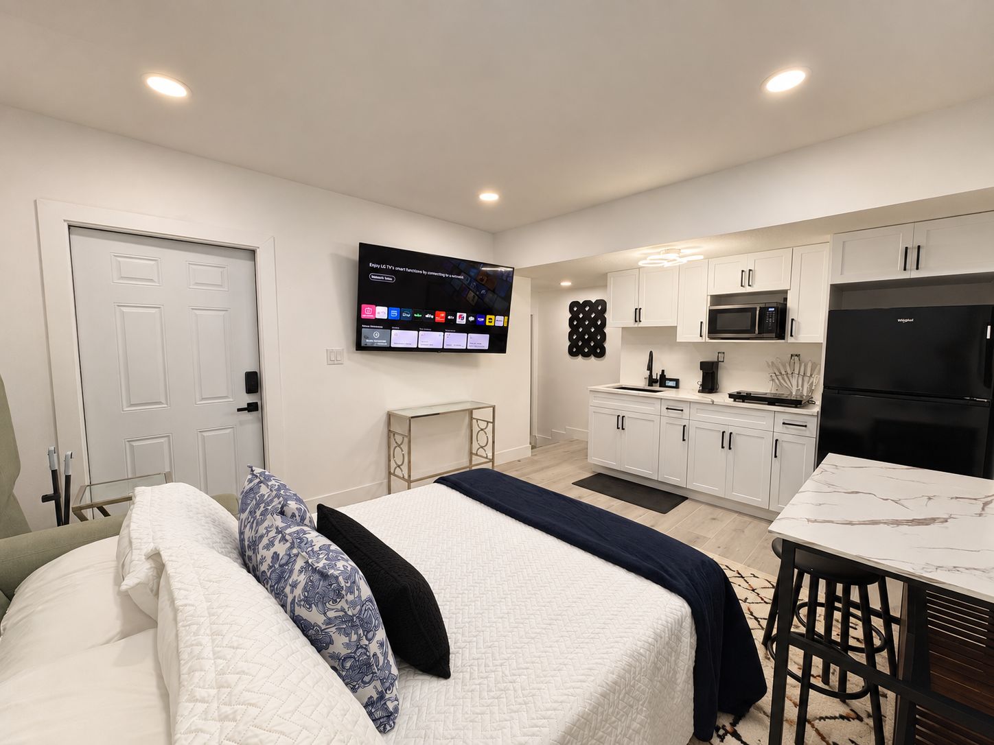 A modern hotel room with a bed, a TV, a kitchenette, and a door.