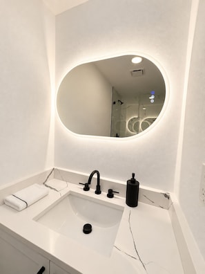 Casa de banho moderna com lavatório branco, torneira preta, dispensador de sabonete e um espelho grande com iluminação integrada.