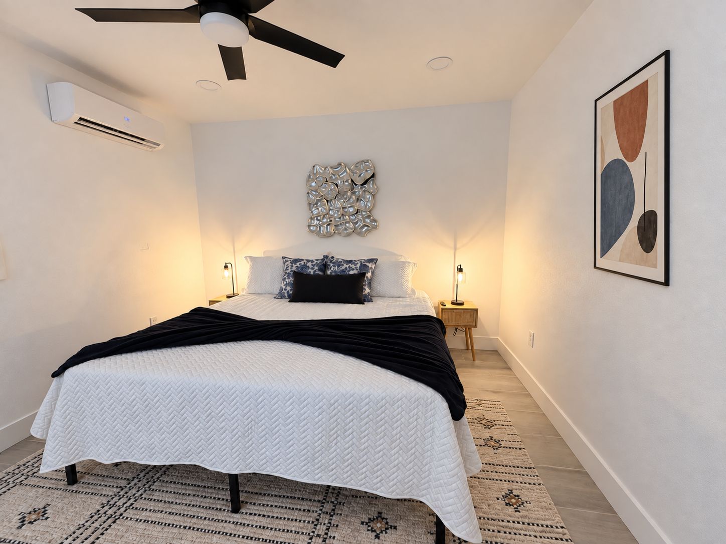 Un dormitorio con cama, ventilador de techo, unidad de aire acondicionado, lámparas de noche, un adorno en la pared y un cuadro abstracto enmarcado.