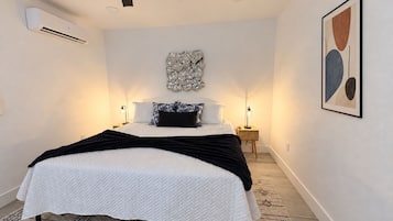 Un dormitorio con cama, ventilador de techo, unidad de aire acondicionado, lámparas de noche, un adorno en la pared y un cuadro abstracto enmarcado.