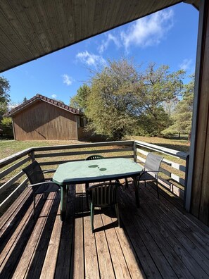 An outdoor wooden deck with a table and chairs, surrounded by a wooden fence, overlooking a grassy area with trees and a building. - LE CHAMOIS No. 8 - A 6-person chalet surrounded by nature, lakes, and waterfalls in the Jura (Uxelles)