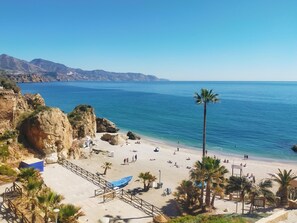 A beach with clear blue water, rocky cliffs, and palm trees. - Nerja Sun Apartments Diputación 8 Center (Nerja)