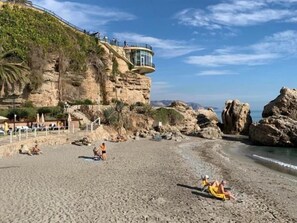 Une plage avec un bâtiment adossé à une falaise, un terrain rocheux et un ciel dégagé.
