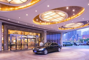 Una entrada de hotel moderna con una puerta giratoria, un coche negro estacionado adelante y vistas de la ciudad a través de grandes ventanales.