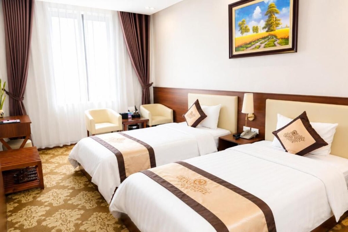 A hotel room with two beds, a desk, a chair, a nightstand, and a painting on the wall.