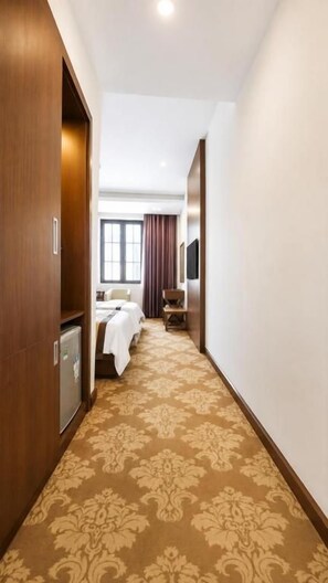 A hotel corridor with a patterned carpet, a bed, a desk, and a television on the wall.