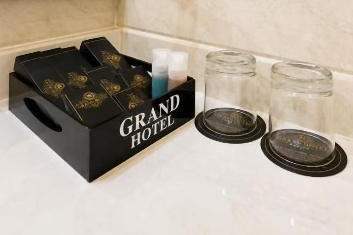 A hotel bathroom with a hairdryer mounted on the wall, a telephone, and a set of toiletries in a black box labeled 'GRAND HOTEL' on the counter.
