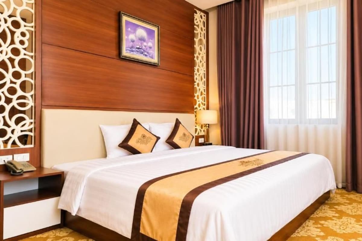 A hotel room with a large bed, a bedside table, a lamp, and a window with curtains.