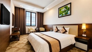 A hotel room with a large bed, a television, a desk, a chair, and a painting on the wall. - Grand Hotel (Hòa Bình)