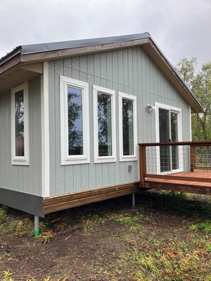 A small, single-story building with a flat roof, white-framed windows, and a wooden deck. - Beautiful new and modern cabin with gorgeous Ninilchik River views. (Ninilchik)