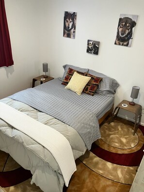 A neatly made bed with a grey and white duvet, a beige throw, and patterned pillows. There are two wall-mounted lamps on nightstands beside the bed, and a round rug with a paw print design. - Beautiful new and modern cabin with gorgeous Ninilchik River views. (Ninilchik)
