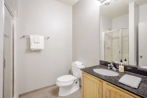 Un baño moderno con cabina de ducha de vidrio, inodoro blanco, lavamanos con encimera de granito y armarios de madera.