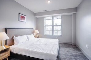 Una habitación de hotel moderna con una cama grande, dos mesitas de noche con lámparas, una ventana con persianas y un cuadro en la pared.