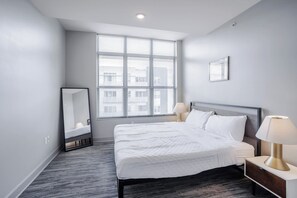 A modern hotel room with a large window, a bed with white bedding, a bedside table with a lamp, and a mirror on the wall.