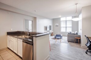A modern kitchen with a dishwasher, a dining area with a sofa and a TV, and a large window providing natural light.