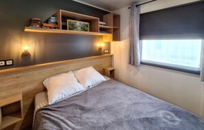 Ein Schlafzimmer mit einem Bett, einem Nachttisch, einem Regal mit Büchern und Snacks sowie einem Fenster mit Vorhängen.