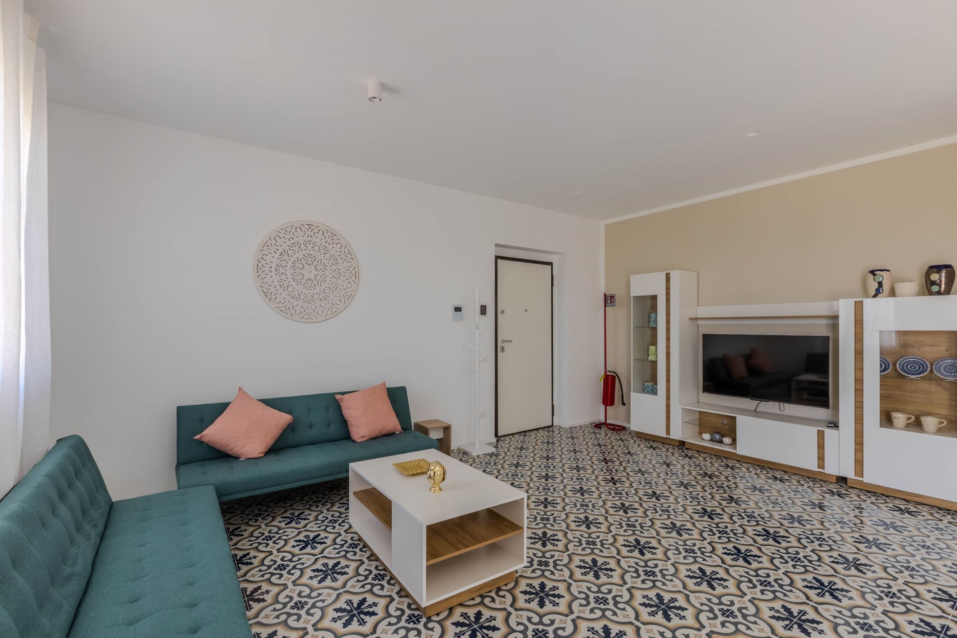Ein Wohnzimmer mit einem salbeengrünfarbenen Sofa, einem weißen Couchtisch, einem Fernseher und fußbodenhohen, gemusterten Fliesen.