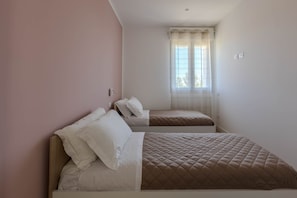 Ein kleines Schlafzimmer mit zwei Betten, einem Fenster mit durchsichtigen Vorhängen und einer schlichten Wand.