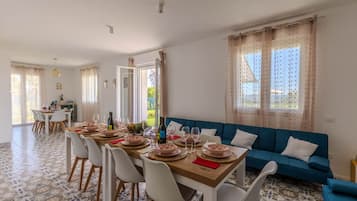 Ein Essbereich mit einem Holztisch, der für sechs Personen gedeckt ist, umgeben von weißen Stühlen. Eine blaue Couch steht an der Wand, und ein Fenster mit Vorhängen ist zu sehen.