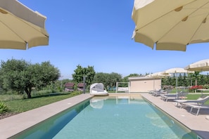 Ein Pool mit Liegestühlen und Sonnenschirmen, umgeben von Grün und blauem Himmel.