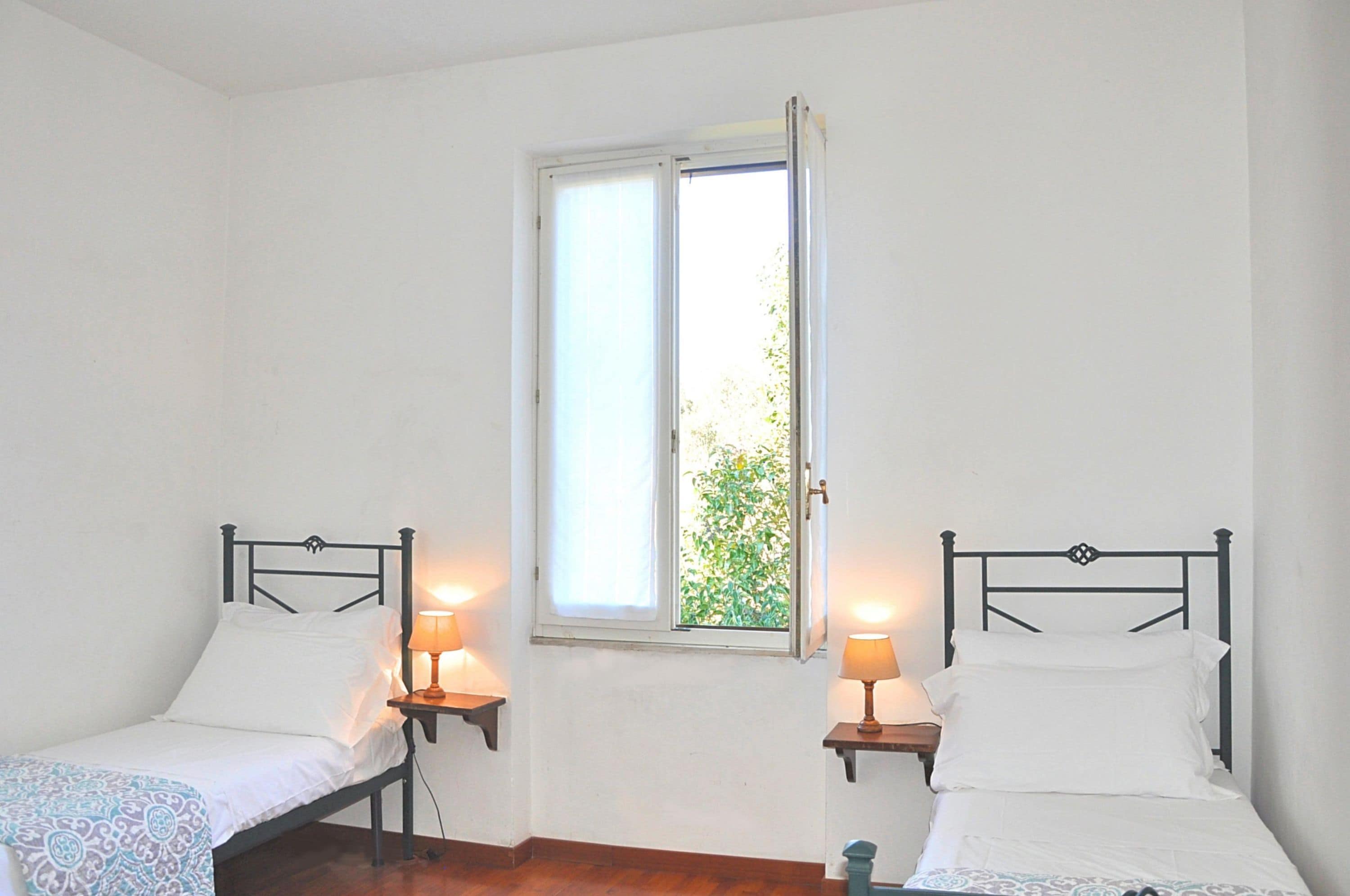 A room with two single beds, each with a bedside table and lamp, a window with a view of greenery, and white walls.