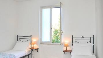 A room with two single beds, each with a bedside table and lamp, a window with a view of greenery, and white walls.