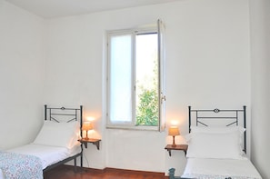A room with two single beds, each with a bedside table and lamp, a window with a view of greenery, and white walls.