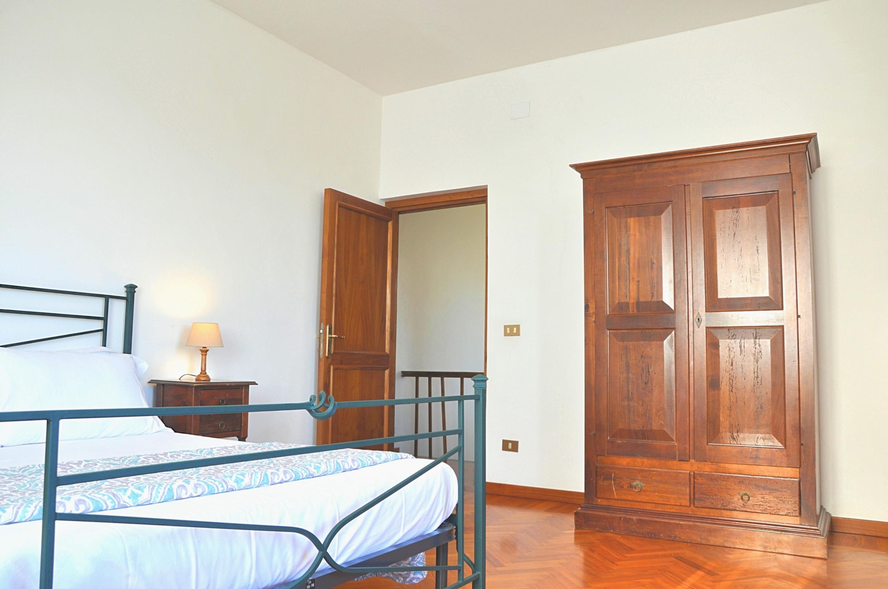 A bedroom with a bed, bedside table, lamp, wardrobe, and a door leading to another room.