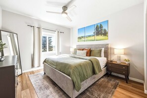 A bedroom with a bed, nightstands, a mirror, a ceiling fan, a window with curtains, and a wall-mounted painting. - The Donnelly Den | Tamarack Getaway, Game Room (Donnelly)