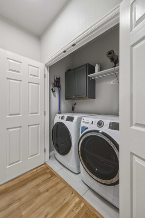 A laundry room with a front-loading washer and dryer, wooden flooring, and a white door. - The Donnelly Den | Tamarack Getaway, Game Room (Donnelly)
