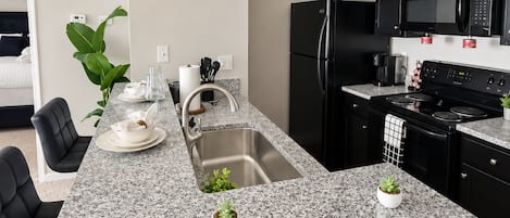 A modern kitchen with a granite countertop, stainless steel sink, black appliances, and a pendant light.